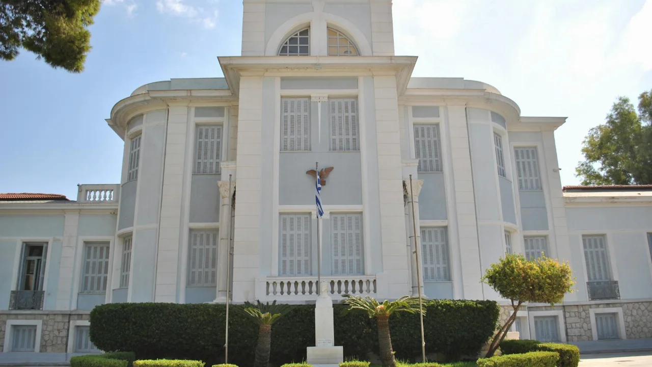 A grand historic building adorned with a Greek flag, surrounded by gardens.