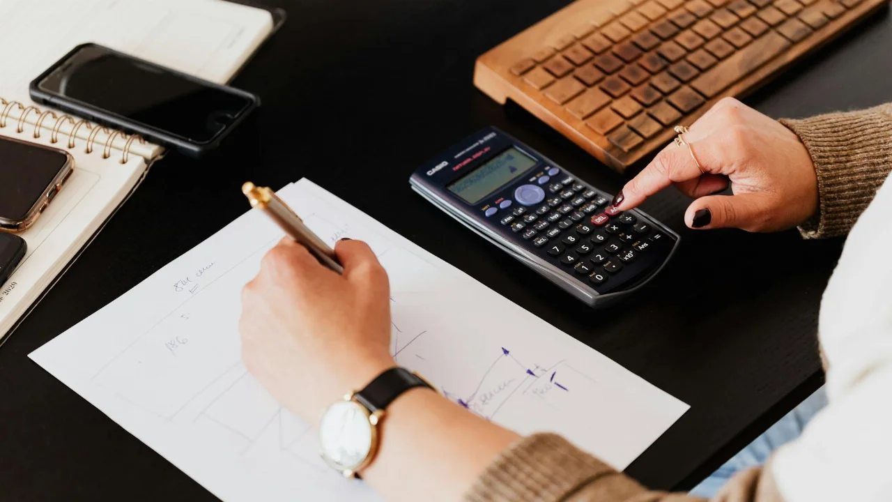 From above crop unrecognizable female accountant wearing stylish watches using calculator and writing down information on paper while working at desk with gadgets